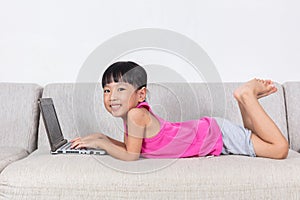 Smiling Asian Chinese little girl laying on sofa using laptop