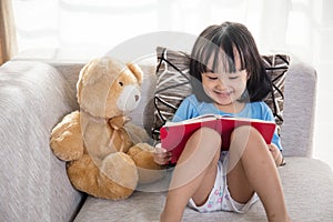 Smiling Asian Chinese little girl reading book with teddy bear