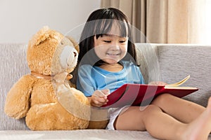 Smiling Asian Chinese little girl reading book with teddy bear