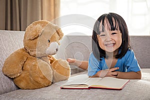 Smiling Asian Chinese little girl reading book with teddy bear