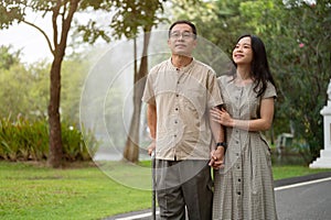 A smiling Asian daughter and her dad with a walking stick are walking in the park together