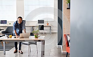 Smiling Asian designer leaning on his desk using a laptop