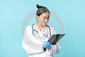 Smiling asian doctor, nurse writing down on clipboard, standing in medical uniform over blue background