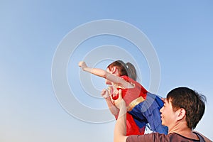 Smiling Asian father lifting up little cute child daughter in superhero dress flying having fun together on blue sky background.