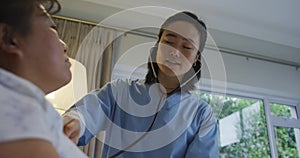 Smiling asian female doctor using stethoscope examining female patient lying in bed at hospital