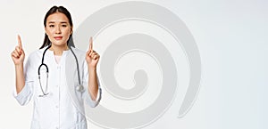 Smiling asian female physician, pointing fingers up and looking at camera, provide medical healthcare information, white