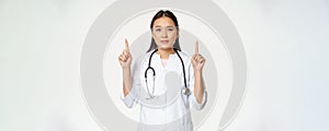 Smiling asian female physician, pointing fingers up and looking at camera, provide medical healthcare information, white