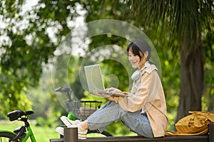 Smiling Asian female student using laptop, sitting on wooden bench in the park