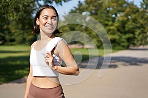 Smiling asian fitness girl holding towel on shoulder, workout in park, sweating after training exercises outdoors