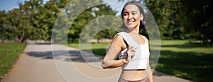 Smiling asian fitness girl holding towel on shoulder, workout in park, sweating after training exercises outdoors