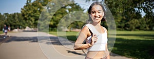 Smiling asian fitness girl holding towel on shoulder, workout in park, sweating after training exercises outdoors