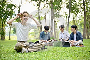 A smiling Asian gay college student sits on the grass in the campus park with his school books