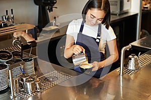 Smiling asian girl barista, cafe staff pouring steamed milk in coffee, prepare cappuccino with latte art, standing in