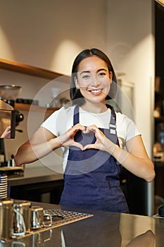 Smiling asian girl barista, shows heart sign, loves making coffee an serving clients, standing in uniform behind counter