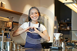 Smiling asian girl barista, shows heart sign, loves making coffee an serving clients, standing in uniform behind counter