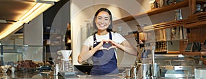 Smiling asian girl barista, shows heart sign, loves making coffee an serving clients, standing in uniform behind counter