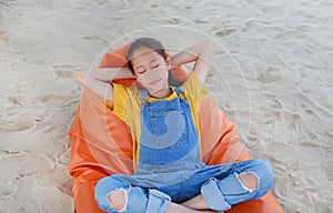 Smiling Asian girl child in dungarees jean relax and sleeping on orange sofa bed beach on sand at summer holiday