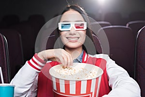 smiling asian girl in 3d glasses with big basket of popcorn watching movie