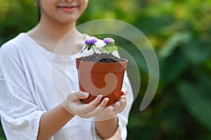 Smiling girl holding potted plant in hands against blurred green nature background on a sunny day. Earth day, Ecology.