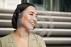 Smiling asian girl, laughing, listening music in headphones, sitting outdoors. Uni student enjoying free time