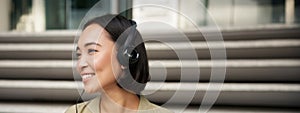 Smiling asian girl, laughing, listening music in headphones, sitting outdoors. Uni student enjoying free time