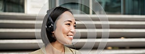 Smiling asian girl, laughing, listening music in headphones, sitting outdoors. Uni student enjoying free time