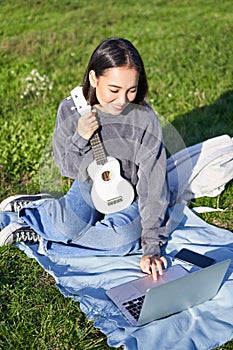Smiling asian girl learns how to play ukulele on laptop, video chat with music teacher, sitting with instrument in park