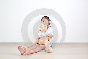 Smiling asian little child girl hugging a teddy bear doll sitting against white wall in the room
