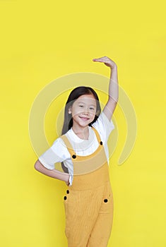 Smiling asian little child girl measures growth by hand isolated over yellow background. Kid estimate her height with looking