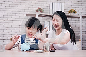 Smiling Asian little asian girl child is putting coins into piggy bank for saving money for the future with mother on wooden table