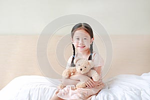 Smiling Asian little girl cuddle teddy bear and looking camera while sitting on the bed at home