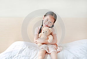 Smiling Asian little girl cuddle teddy bear sitting on the bed at home