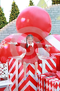 Smiling Asian little girl in red dress sitting on big gift box with arms open wide to present in winter season and happy new year