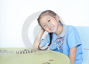 Smiling Asian little girl in school uniform sitting on table with pile of coins for saving over white background. Kid counting