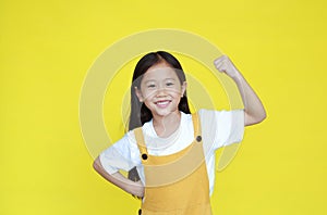 Smiling asian little girl showing his muscle with looking camera isolated on yellow background. Strong kid in dungarees with good