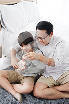 Smiling Asian little son child is putting coins into piggy bank for saving money for the future with father in bedroom. Child