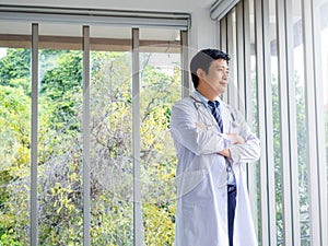 Smiling Asian man doctor portrait in white coat standing alone with crossed arms near glass windows in medical office.