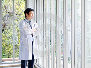 Smiling Asian man doctor portrait in white coat standing alone with crossed arms near glass windows in medical office.