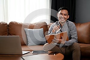 Smiling Asian man making list or taking notes on his notebook while sitting on sofa