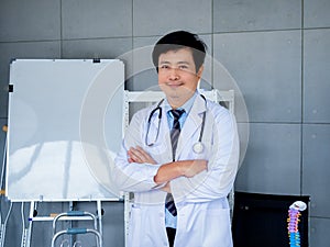 Smiling Asian man orthopedic doctor portrait in white coat standing with crossed arms near white board in medical office.