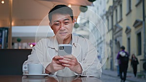 Smiling asian man tourist sitting in street cafe holding mobile phone booking tickets online remotely communicates with