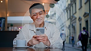 Smiling asian man tourist sitting in street cafe holding mobile phone booking tickets online remotely communicates with
