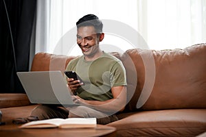 Smiling Asian man using his smartphone and laptop while sitting on a sofa in his living room