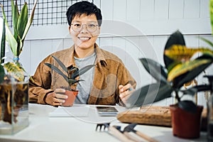 Smiling Asian man in vintage glasses sitting  taking care of the plant  and writing report book at home