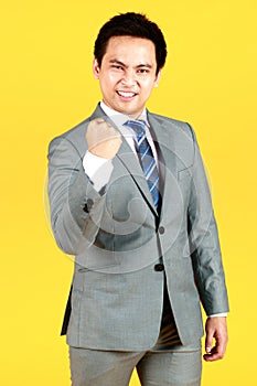 Smiling Asian man wearing a gray suit holding hand raise a fist in yellow background. A businessman personality and well dressed