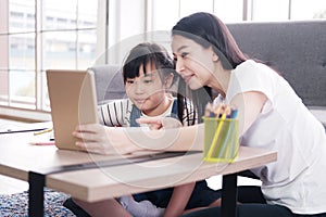 Smiling Asian mother and little girl child is is online learning with laptop together in living room. Homeschool and educational