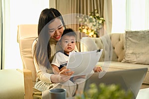 Smiling Asian mother working at home with laptop and documents sitting on armchair with cute little baby son