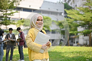 Smiling Asian Muslim female college student carrying her laptop and standing in the campus park