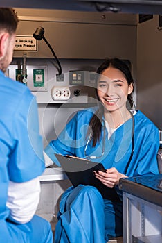 smiling Asian paramedic sitting in ambulance and looking