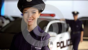 Smiling asian policewoman posing to camera against partner near patrol car, duty